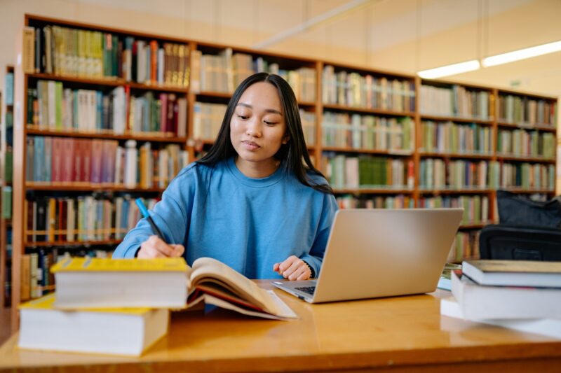 Jovem moça estudando para o Enem dentro de biblioteca, com livros e notebook sobre a mesa