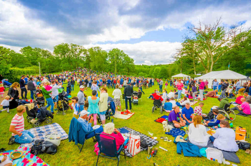 Celebração do solstício de verão, uma das principais festas juninas do mundo, na Suécia. Em local externo com grama e árvores, e muitas roupas coloridas, mantos de piqueniques e guarda-sóis