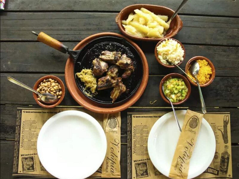 Vasilhas, potes e frigideira com costela de bode na Cachaça e outros alimentos prontos para serem servidos em dois pratos brancos sobre a mesa.
