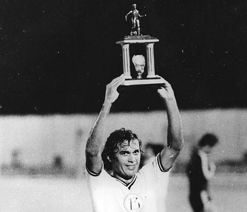 Foto em preto e branco, com jogador Ardelino (que está com os cabelos molhados e sorrindo) com camisa branca do Treze Futebol Clube levantando um troféu