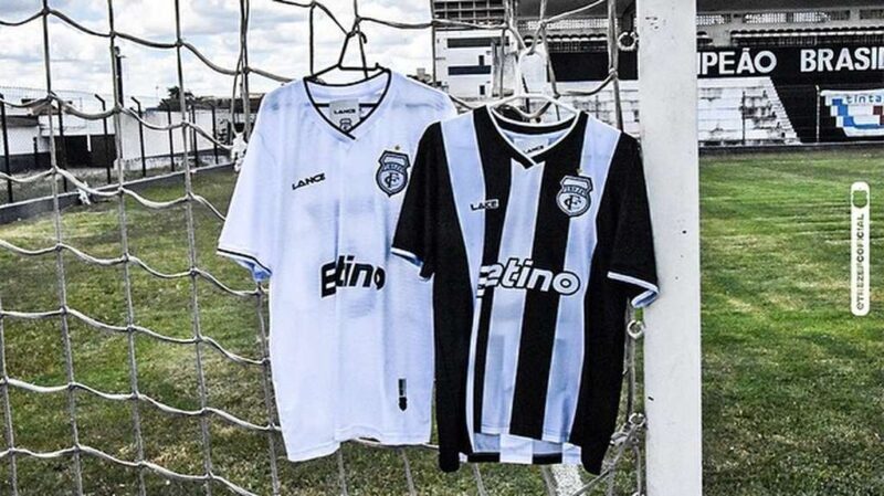 Uniforme do treze FC: uma camisa branca pendurada na rede do gol e, pendurada ao lado, uma camisa com listras verticais em branco e preto